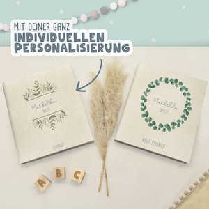 May include: Two beige fabric-covered folders with personalized text and green botanical illustrations. The left folder features the name Mathilda Maier and the word Zeugnisse. The right folder shows a green leaf wreath, the name Mathilda Maier, and the text Meine Zeugnisse. A blue arrow points to the left folder.