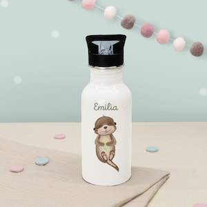 Könnte beinhalten: Weiße Trinkflasche mit schwarzem Deckel. Die Flasche zeigt eine Cartoon-Otter-Illustration und den Namen "Emilia" in grüner Schrift. Im Hintergrund befinden sich pastellfarbene Dekorationen.