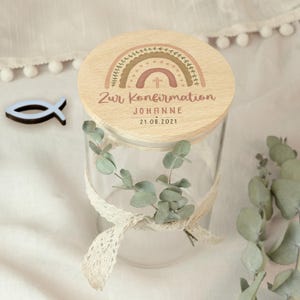 May include: A clear glass jar with a wooden lid featuring a rainbow design and the text "Zur Konfirmation" and "Johanne 21.08.2021". The jar is decorated with a lace ribbon and eucalyptus leaves. A blue fish-shaped wooden cutout is next to the jar.