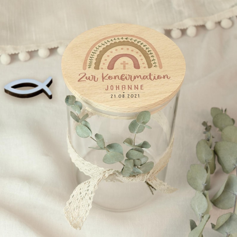 May include: A clear glass jar with a wooden lid featuring a rainbow design and the text "Zur Konfirmation" and "Johanne 21.08.2021". The jar is decorated with a lace ribbon and eucalyptus leaves. A blue fish-shaped wooden cutout is next to the jar.