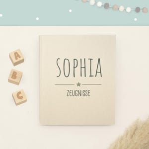May include: A beige rectangular book with the name SOPHIA printed in dark green capital letters, followed by a horizontal line with a small star in the center and the word ZEUGNISSE below it. Three wooden blocks with letters A, B, and C are nearby.