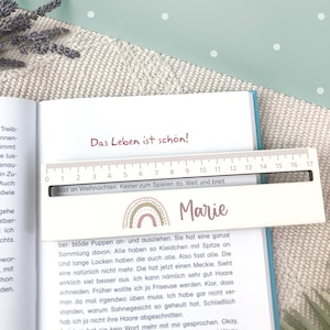May include: A pink and white wooden ruler with a rainbow design and the name "Marie" written on it. The ruler has numbers from 0 to 17 and the text "Das Leben ist sch&ouml;n!" and "bad an Weihnachten. Keiner zum Spielen da. Weit und breit" printed on it.
