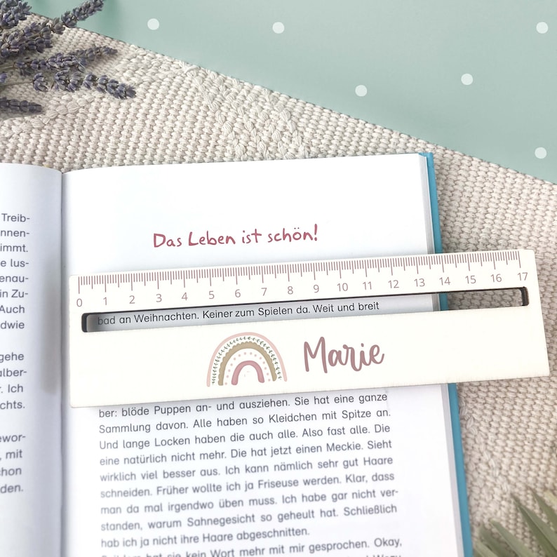 May include: A pink and white wooden ruler with a rainbow design and the name "Marie" written on it. The ruler has numbers from 0 to 17 and the text "Das Leben ist sch&ouml;n!" and "bad an Weihnachten. Keiner zum Spielen da. Weit und breit" printed on it.