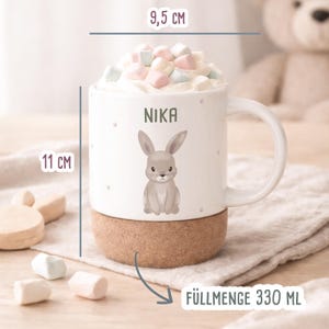 May include: A white ceramic mug with a cork base, 4.3 inches tall and 3.7 inches wide, featuring a gray bunny illustration and the name "NIKA". The mug is filled with marshmallows and has a capacity of 11.1 fluid ounces.