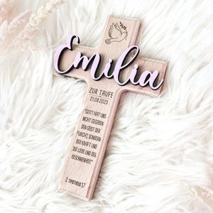 May include: Wooden cross with a pink "Emilia" inscription, a dove design, and the text "Zur Taufe 21.08.2023" and a quote about faith.