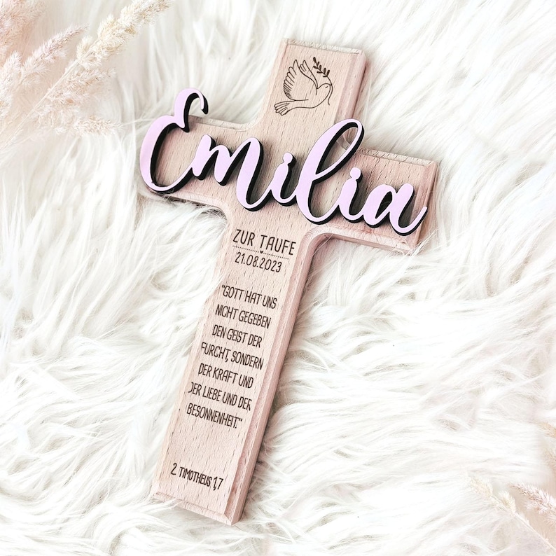 May include: Wooden cross with a pink "Emilia" inscription, a dove design, and the text "Zur Taufe 21.08.2023" and a quote about faith.