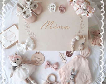 May include: A wooden box with the name "Mina" engraved on the lid. The box is filled with baby items, including a plush toy, a rattle, a pacifier, a headband, and a blanket. The box is surrounded by a white lace trim.