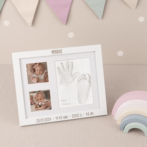 May include: White frame with a white mount and two photographs of a baby girl. The top photo shows the baby wearing a pink outfit and holding a soft toy. The bottom photo shows the baby wearing a white outfit and holding a soft toy. The frame also includes a handprint and footprint of the baby. The frame has the baby's name "MARIE" and the date and time of birth, "23.01.2024 - 11:45 UHR - 3500 G - 46 CM".