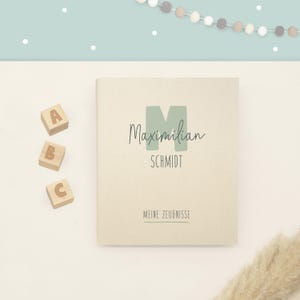 May include: A cream-colored rectangular book with a large sage green letter M, the name Maximilian Schmidt, and the text MEINE ZEUGNISSE printed on the cover. Three small wooden blocks with the letters A, B, and C are placed to the left.