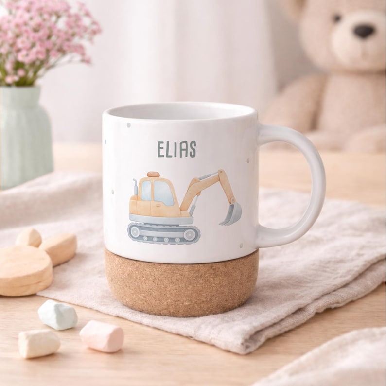 May include: White ceramic mug with a cartoon excavator design and the name "ELIAS" printed on it. The mug sits on a cork base. The excavator is yellow and beige with a gray bucket. Small gray dots are scattered around the mug.