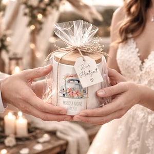 Könnte beinhalten: Ein weißes Keramikglas mit Holzdeckel, verpackt in durchsichtigem Zellophan und mit Bindfaden verschnürt. Das Glas zeigt ein Blumendesign mit einem Oldtimer und den Namen "Moritz & Nadine" und dem Datum "20.02.2021". Ein Anhänger lautet "Zur Hochzeit".