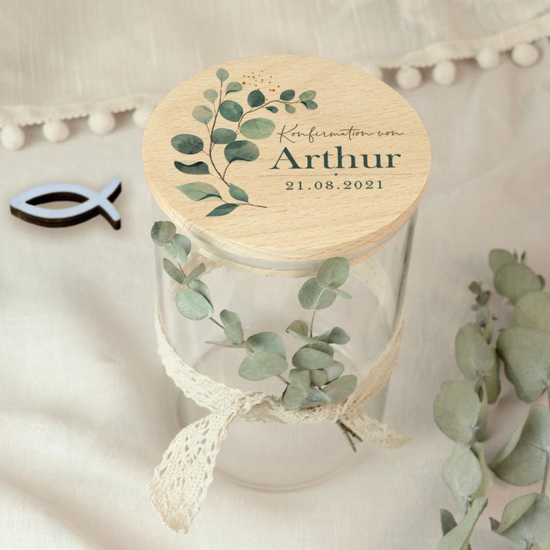 May include: A clear glass jar with a wooden lid featuring a botanical design and the text "Konfirmation von Arthur 21.08.2021". The jar is adorned with a lace ribbon and eucalyptus leaves. A small blue fish-shaped decoration is also present.