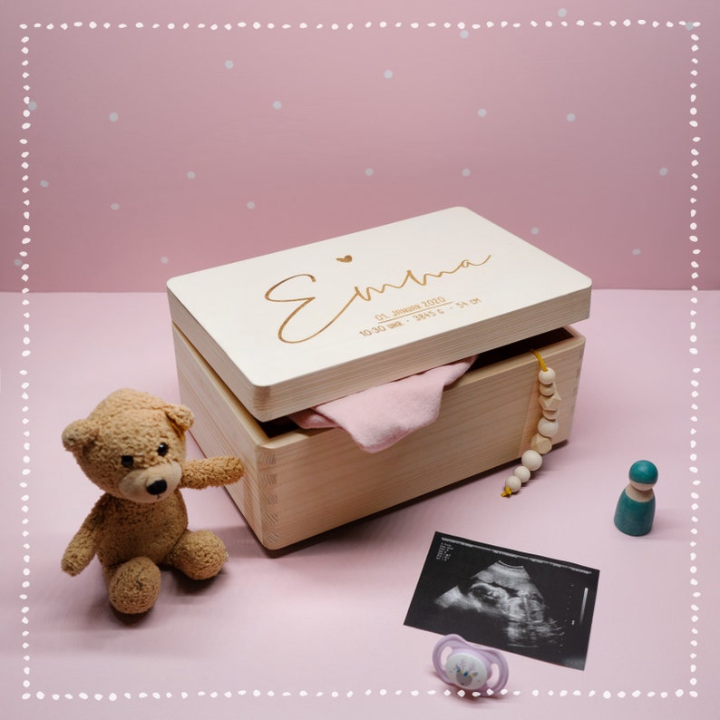 Pu&ograve; includere: Una scatola di legno per ricordi con una finitura in legno chiaro e un'iscrizione dorata che recita "Emma 01 gennaio 2020 10:30 AM - 3,940 g - 54 cm". La scatola &egrave; aperta e contiene un body rosa per neonato, una collana di perline di legno e una piccola bambola di legno.