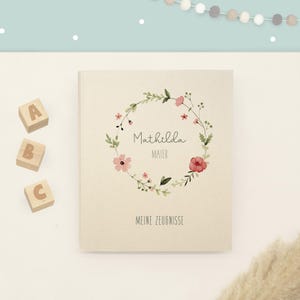 May include: A cream-coloured hardcover book featuring a watercolour floral wreath illustration surrounding the printed text Mathilda Maier and the words MEINE ZEUGNISSE at the bottom, accompanied by three wooden blocks labelled A, B, and C.