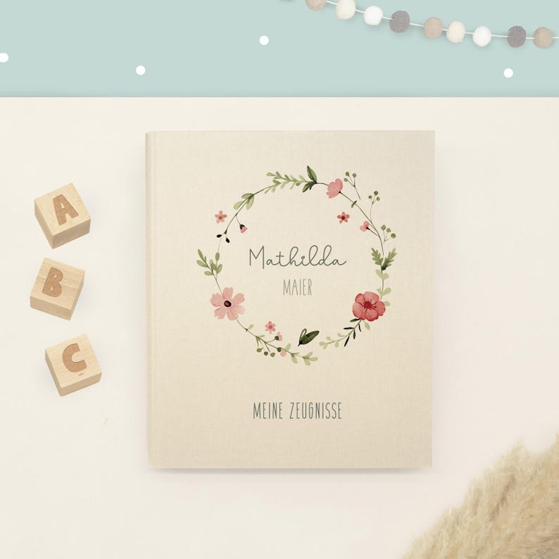 May include: A cream-coloured hardcover book featuring a watercolour floral wreath illustration surrounding the printed text Mathilda Maier and the words MEINE ZEUGNISSE at the bottom, accompanied by three wooden blocks labelled A, B, and C.