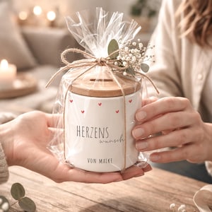 Könnte beinhalten: Ein weißer Keramik-Sparschwein mit Holzdeckel, verziert mit kleinen roten Herzen. Das Sparschwein ist in durchsichtiges Zellophan gewickelt und mit Bindfaden verschnürt, mit dem Text "HERZENS wunsch VON MAIKE".