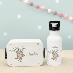 Könnte beinhalten: Weißes Lunchbox- und Trinkflaschen-Set. Beide Artikel zeigen ein Cartoon-Affen-Design mit dem Namen "Emilia". Die Lunchbox hat schwarze Verschlüsse, und die Trinkflasche hat einen schwarzen Deckel.