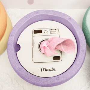 Può includere: Un contenitore rotondo color lavanda con un coperchio bianco con un'illustrazione di lavatrice e il nome "Moritz". Un panno rosa è parzialmente visibile. Il contenitore è progettato per il bucato o lo stoccaggio.