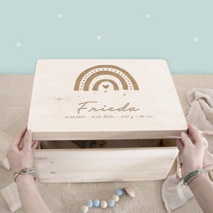 Op de afbeelding: Een houten opbergdoos met een regenboogontwerp en de naam "Frieda" gegraveerd op de deksel. De doos bevat ook de datum "16.05.2023", de tijd "21:28 Uhr", het gewicht "3100 g" en de lengte "50 cm".