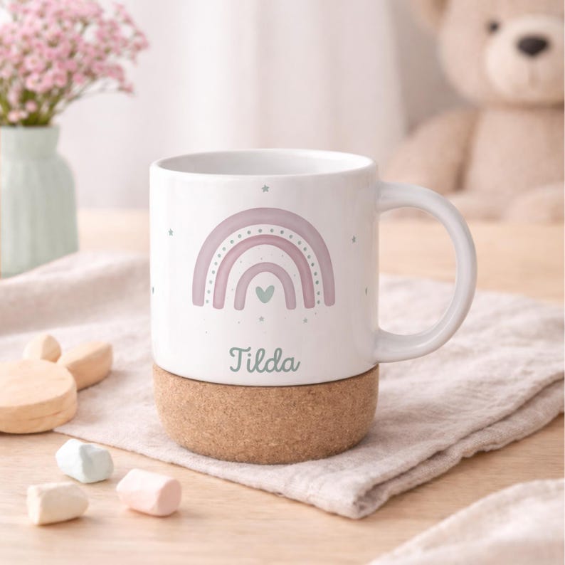 May include: White ceramic mug with a pastel rainbow design, a green heart, and the name "Tilda" in a script font. The mug sits on a cork base. Stars are scattered around the rainbow. A perfect gift for a child.