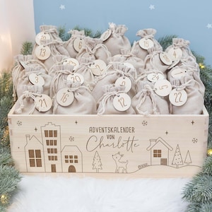 Op de afbeelding: Een houten adventskalenderbox gevuld met kleine beige stoffen zakjes, elk genummerd met een houten label. De doos is versierd met geëtste huis- en boomontwerpen en de tekst "ADVENTSKALENDER von Charlotte".