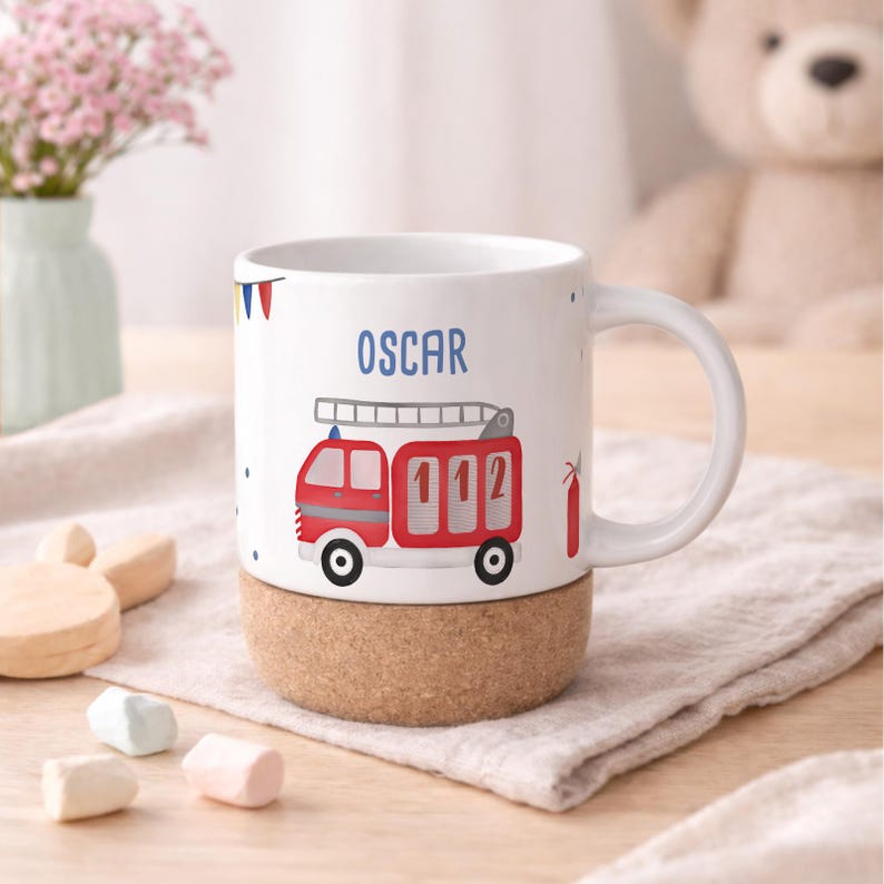 May include: White ceramic mug with a red fire truck design, the number 112, and the name "OSCAR" in blue. The base is made of cork. A small red fire extinguisher is also visible.
