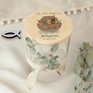 May include: A clear glass jar with a wooden lid featuring an illustration of Noah's Ark with animals. The lid has the text "ZUR KONFIRMATION", the name "Johann", and the date "21.08.2021". A white lace ribbon and eucalyptus sprigs decorate the jar.