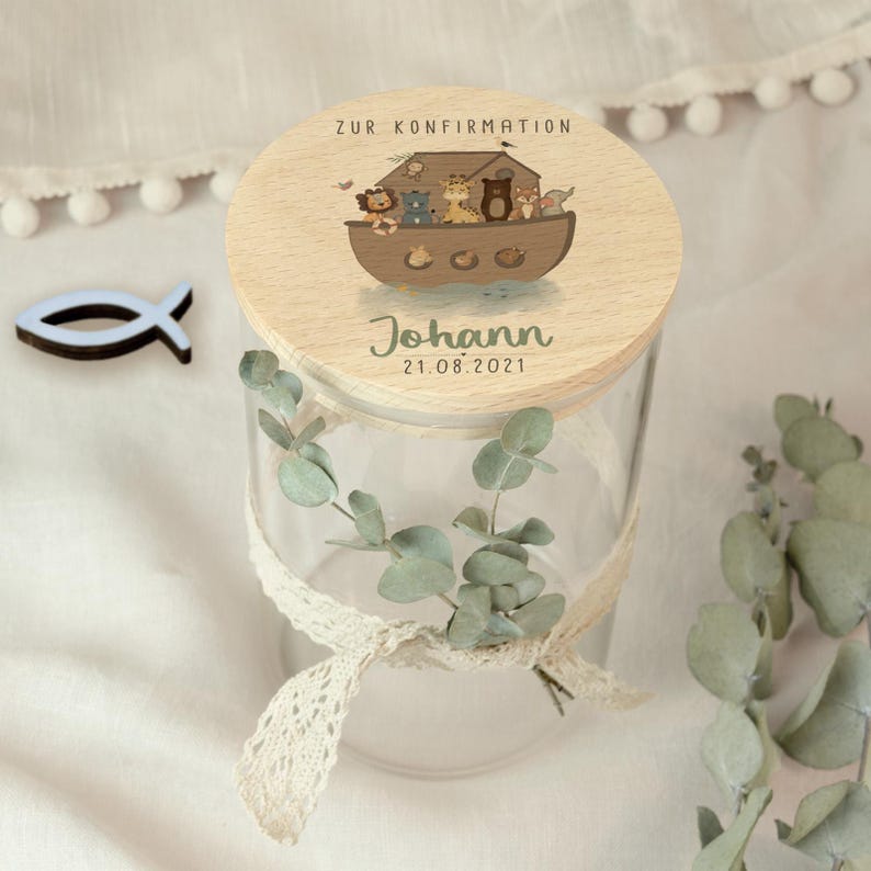 May include: A clear glass jar with a wooden lid featuring an illustration of Noah's Ark with animals. The lid has the text "ZUR KONFIRMATION", the name "Johann", and the date "21.08.2021". A white lace ribbon and eucalyptus sprigs decorate the jar.