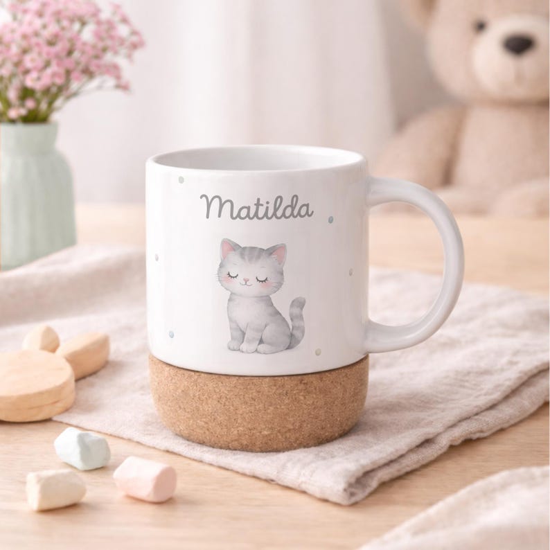 May include: White ceramic mug with a gray cat illustration and the name "Matilda" printed in gray. The mug sits on a cork base. The mug has a handle and is decorated with small gray dots.