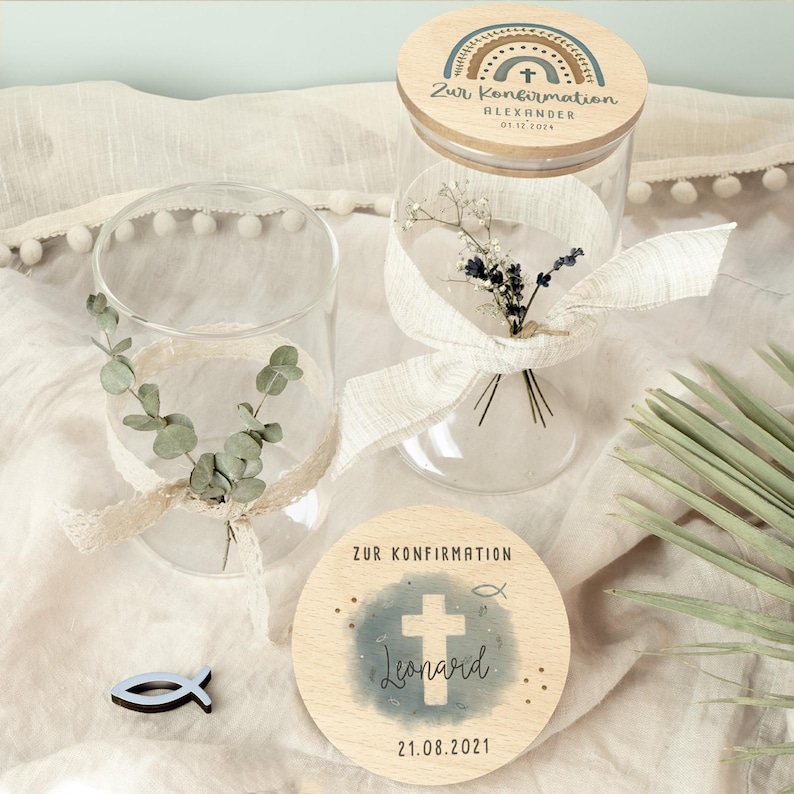 May include: Two clear glass jars with wooden lids and a small wooden fish. One jar has a bouquet of dried flowers and a ribbon. The other jar has eucalyptus leaves and a ribbon. The lids and a wooden disc are decorated with text and a cross.