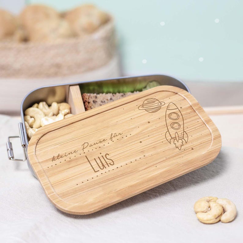 May include: A silver lunchbox with a wooden lid. The lid has a rocket and planet design with the text "kleine Pause f&uuml;r Luis".