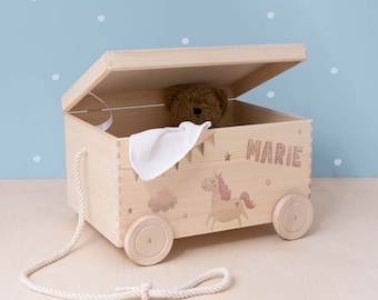 May include: A wooden toy chest with wheels and a pull rope. The chest is decorated with a unicorn, clouds, and stars. The name "MARIE" is written on the side of the chest.