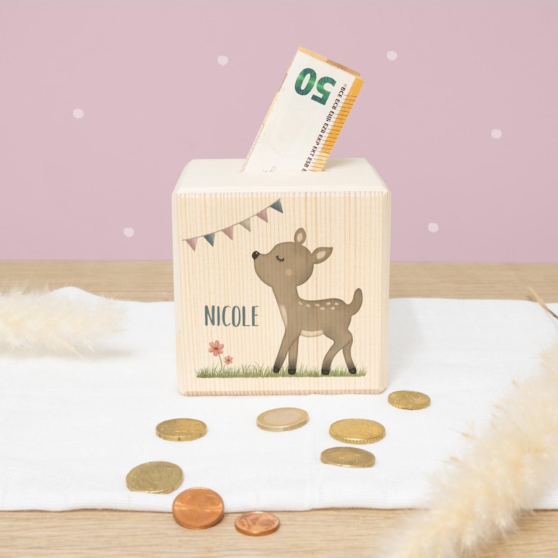 May include: A wooden piggy bank shaped like a cube with a slot on the top. The front of the bank features a cartoon illustration of a fawn with the name "NICOLE" printed below. The bank is sitting on a white surface with a few coins scattered around it.