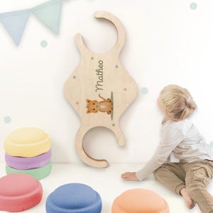Può includere: Un gancio da parete in legno chiaro, dalla forma unica, con il nome "Matheo" e un'illustrazione di un gatto dei cartoni animati. Il gancio ha un design curvo e piccoli fori circolari. Elementi decorativi dai colori pastello sullo sfondo.