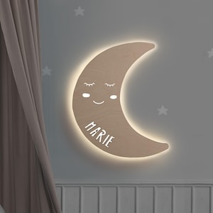 May include: A crescent moon-shaped wall light made of light-toned wood. The moon features a sleepy face with closed eyes, a small smile, and rosy cheeks. The name MARIE is printed in white capital letters. The light emits a soft glow from behind.