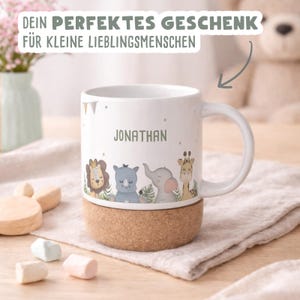May include: White ceramic mug with a cork base, featuring a cartoon safari animal design and the name "JONATHAN" in green. The mug has a handle and is decorated with small flags. Text at the top reads "DEIN PERFEKTES GESCHENK F&Uuml;R KLEINE LIEBLINGSMENSCHEN".