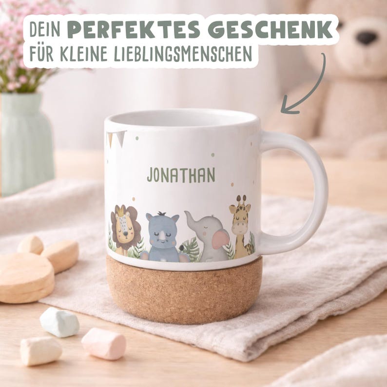 May include: White ceramic mug with a cork base, featuring a cartoon safari animal design and the name "JONATHAN" in green. The mug has a handle and is decorated with small flags. Text at the top reads "DEIN PERFEKTES GESCHENK F&Uuml;R KLEINE LIEBLINGSMENSCHEN".