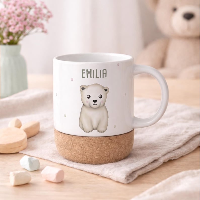 May include: White ceramic mug with a polar bear illustration and the name "EMILIA" printed on it. The mug has a cork base and a white handle. The background includes a vase of pink flowers and a teddy bear.