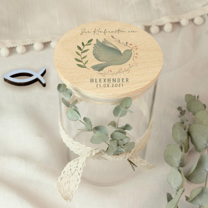 May include: A clear glass jar with a wooden lid featuring a dove illustration and the text "Zur Konfirmation von ALEXANDER 21.08.2021". The jar is decorated with a lace ribbon and eucalyptus leaves. A small blue fish-shaped ornament is next to the jar.