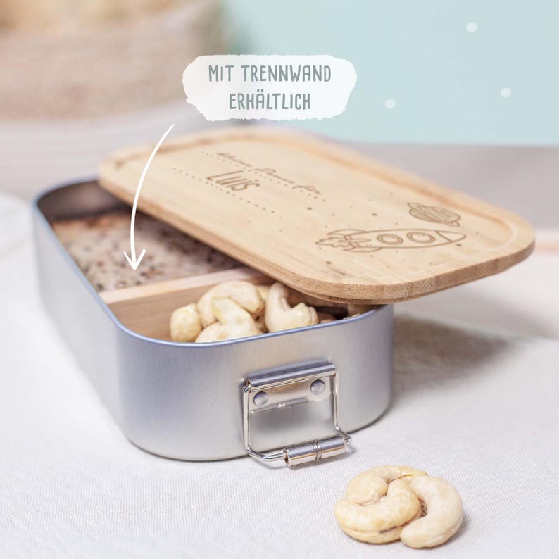 May include: A silver lunchbox with a wooden lid featuring a rocket and planets design. The lid is engraved with the name "Lilis". The lunchbox has a divider inside and is filled with cashews.