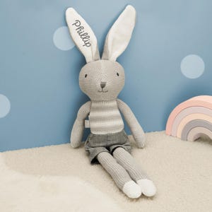 Könnte beinhalten: Ein gestricktes Stoffhasen-Spielzeug mit langen Ohren, einem grau-weiß gestreiften Oberkörper und einer grauen Hose. Der Name Phillip ist in schwarzer Schreibschrift auf das linke Innenohr gestickt. Der Hase hat einen grauen Kopf, lange Arme und Beine.