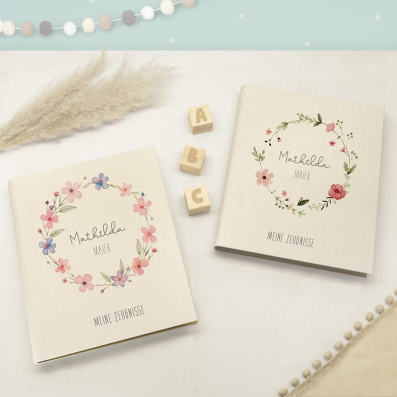 May include: Two off-white fabric-covered folders featuring a watercolour floral wreath design. Each folder displays the name Mathilda Maier and the text MEINE ZEUGNISSE. Three small wooden blocks with the letters A, B, and C sit between them.