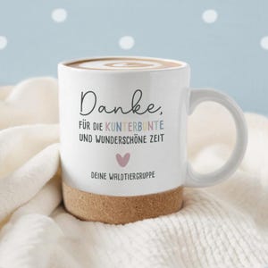 May include: White ceramic mug with a cork base, filled with a coffee drink. The mug has a curved handle and features the text "Danke" and other German text in black, blue, and pink. A pink heart is below the text.