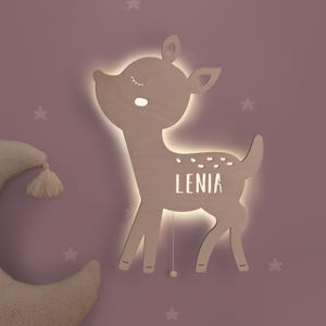 May include: A light brown wooden wall lamp shaped like a deer with a glowing backlight. The deer has closed eyes, a small mouth, and white spots on its back. The name LENIA is cut out from the centre of the deer, allowing light to shine through.