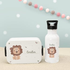 Könnte beinhalten: Weißes Lunchbox- und Trinkflaschen-Set. Beide Artikel zeigen eine Cartoon-Löwen-Illustration und den Namen "Emilia". Die Lunchbox hat schwarze Verschlüsse und die Wasserflasche einen schwarzen Deckel. Im Hintergrund hängt eine Girlande.
