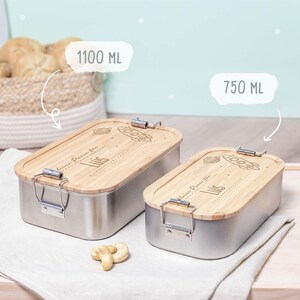 May include: Two stainless steel lunch boxes with bamboo lids. The lids have a rocket ship and planet design with the name "Luis" engraved on them. The larger lunch box is labeled "1100 ML" and the smaller lunch box is labeled "750 ML".
