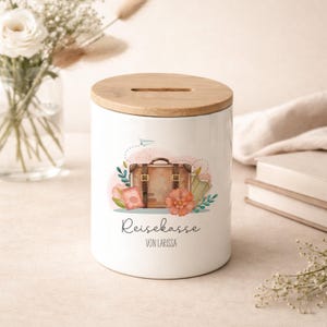 Può includere: Salvadanai in ceramica bianca con coperchio in legno. La banca presenta un'illustrazione ad acquerello di una valigia, un passaporto e un fiore. Il testo "Reisekasse VON LARISSA" è scritto sulla banca.
