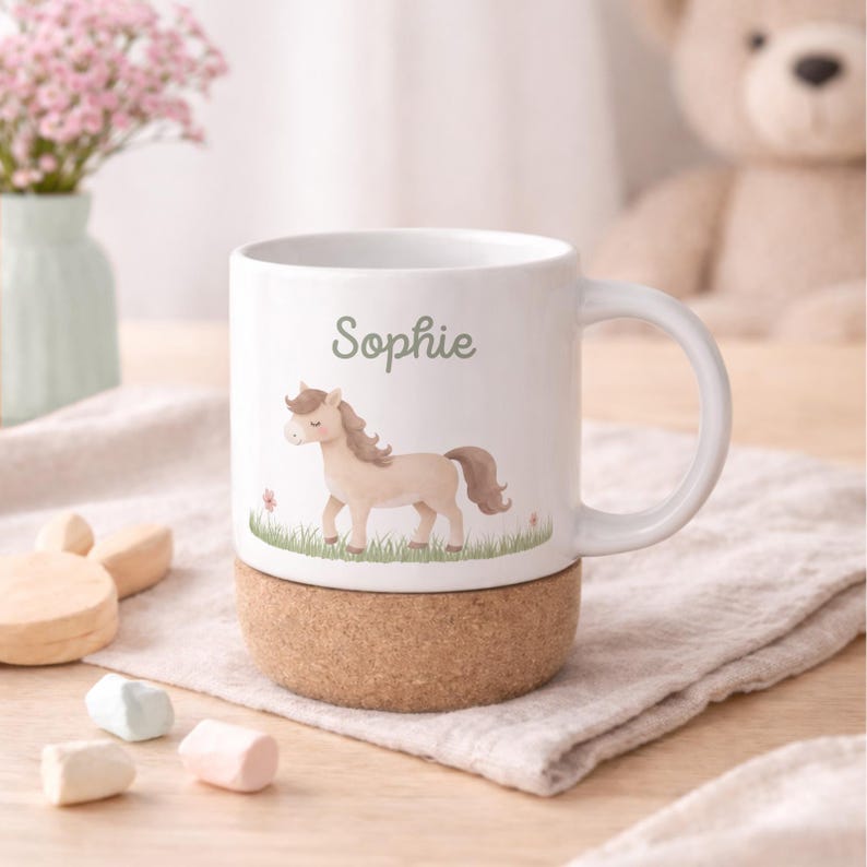 May include: White ceramic mug with a cork base, featuring a watercolor illustration of a tan pony and the name "Sophie" in green. The mug has a curved handle and is set on a light-colored surface.