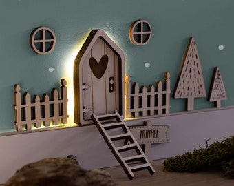 May include: A wooden fairy door with a heart-shaped window, a ladder, and a fence. The door is lit up with a warm light and the sign reads "Mumpel".