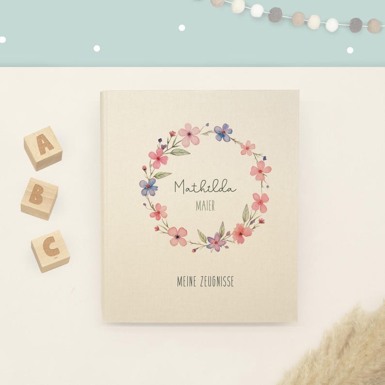 May include: A beige hardcover book featuring a watercolour floral wreath in pink, blue, and green hues. The text reads Mathilda Maier and MEINE ZEUGNISSE. Three wooden blocks with the letters A, B, and C are positioned to the left of the book.